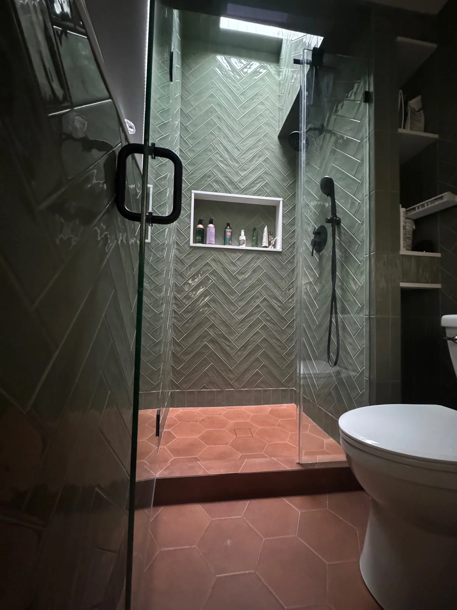 Walk-in shower in glossy green herringbone tile with a terracotta hexagon floor